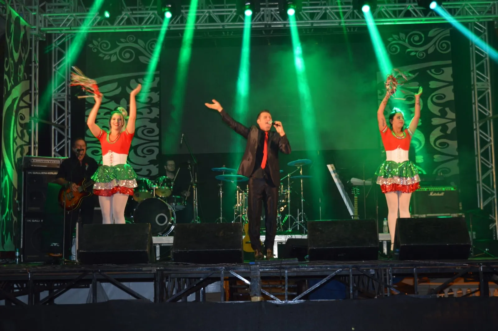 Palco da festa iluminado em verde, em edição anterior: cantor de terno preto no centro e duas dançarinas em trajes com as cores da Itália